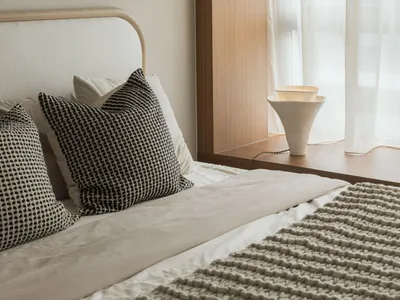 A minimalist bedroom with white bedding, cushions, and a lamp.
