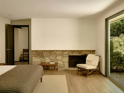 A minimalist bedroom featuring a bed with a wooden headboard, a stone fireplace, and a lounge chair near a large window.
