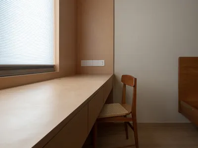 A minimalist home office features a wooden counter and chair against a white wall, with a window allowing natural light in.