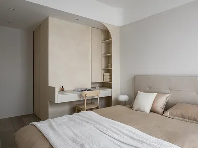 Modern, minimalist bedroom with a beige headboard bed, wooden floor, and built-in desk with shelves.