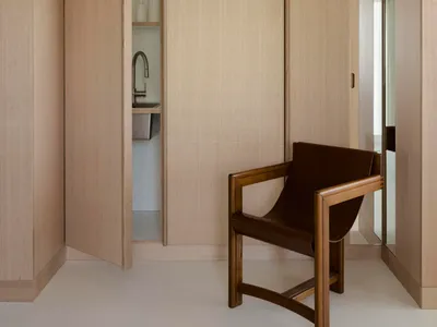 A minimalist bedroom with wooden walls and floor, featuring a chair in the foreground and a small kitchen area in the backgro