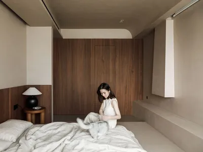 A woman sitting on a bed, petting a cat, with a bedside lamp.