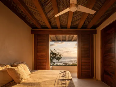 A cozy bedroom featuring wooden beams on the ceiling, a bed with fabric bedding, and a coastal view through the window.