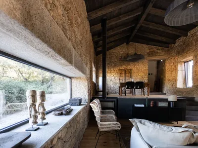 An industrial-style living room with stone walls, a rustic wooden ceiling, and a large window overlooking the outdoors.