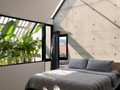 A minimalist bedroom featuring concrete walls and large windows with green plants.