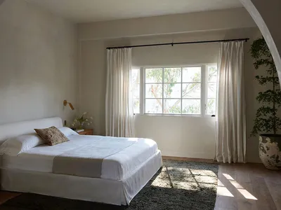 A minimalist bedroom featuring a white fabric bed with a plain white headboard, white bedding, and a natural light streaming