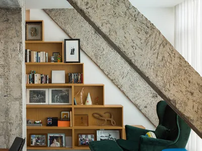 A modern industrial living room featuring a wooden bookshelf and concrete staircase, with a green velvet armchair and ottoman