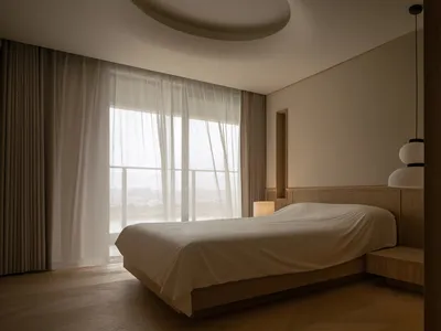 Modern bedroom with a neutral color palette, featuring a bed with a wooden frame and a large window with curtains.