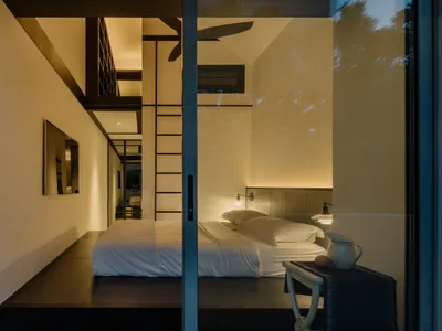 A modern minimalist bedroom with a wooden bed platform and white bedding, illuminated by soft lighting.