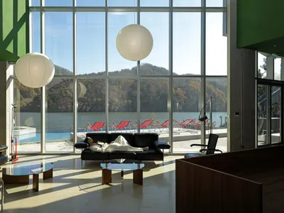 A modern hotel lobby features a glass wall overlooking a serene mountain lake, accompanied by black leather couches and red l