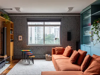 A spacious, industrial-style living room with brick and concrete walls, an orange sectional sofa, a large window, and a mix o