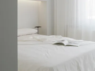 A minimalist bedroom with white walls, curtains, and bedding, featuring a lighted alcove above the bed.
