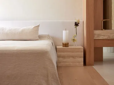 Minimalist bedroom with a wooden floor, beige bedding, and a wooden nightstand with a candle and a vase of flowers.