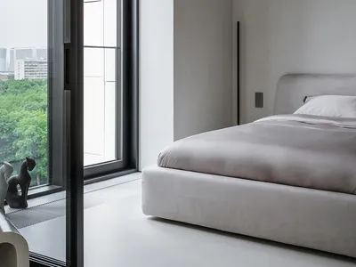 Modern minimalist bedroom featuring a grey upholstered bed with a fabric headboard, large window with urban view, and sleek d