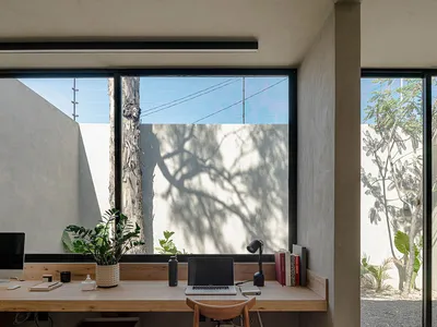A modern home office featuring a wooden desk with a laptop, lamp, and books, set against a backdrop of large windows showcasi