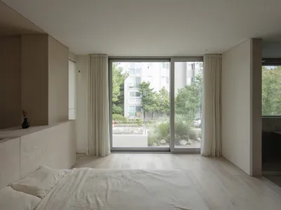 A minimalist bedroom featuring neutral colors, a large window with white curtains, and a view of trees and buildings outside.