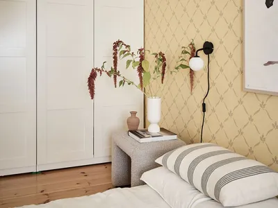 Minimalist bedroom with neutral tones featuring a bed, pillows, and a small bench with a white vase and flowers.