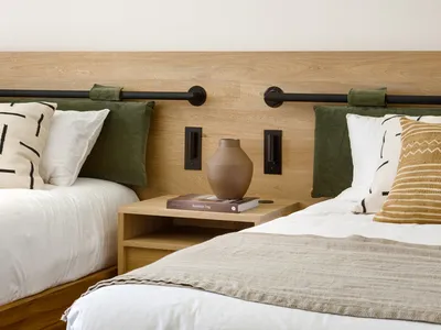 Minimalist bedroom features a bed with a wooden headboard, beige and white bedding, and a side table with a beige vase and bo