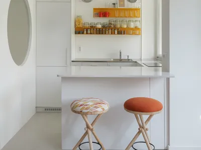 A minimalist kitchen with white cabinets, a white countertop, and colorful stools.