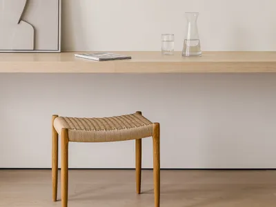 A minimalist home office desk with a wooden top, a rope stool, a framed abstract artwork, and a glass carafe with a glass of