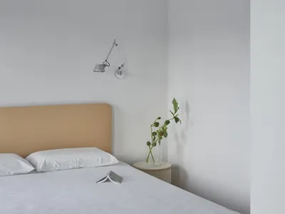 A modern minimalist bedroom featuring a bed with a fabric headboard and white bedding, placed in front of a white wall with a