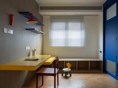 A minimalist home office with wooden furniture, including a yellow desk, a red wall lamp, and a child's chair.