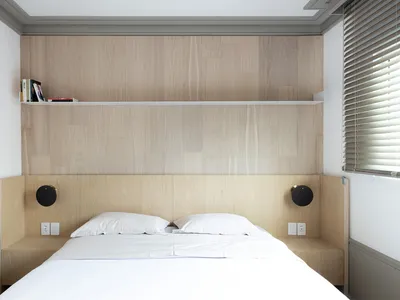 A minimalistic bedroom with wooden walls and a white bed, featuring a built-in shelf above the headboard.