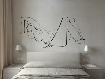 A minimalist bedroom featuring a bed with a white headboard and linens, a wall drawing of a reclining figure, and bedside lam