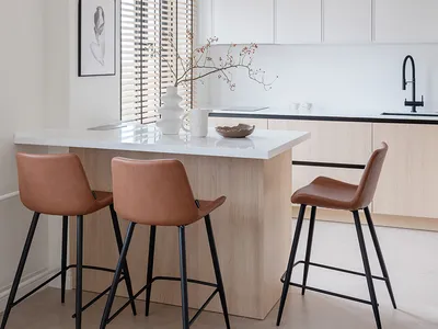 A modern minimalist kitchen with wooden finishes, a hanging woven ceiling lamp, and brown leather bar stools.