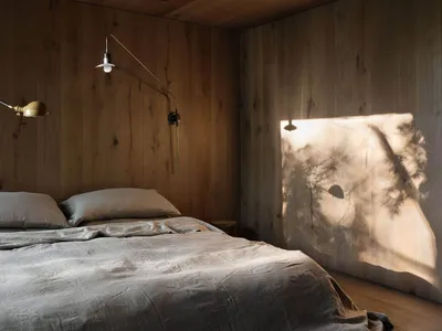 A minimalist bedroom with wooden walls and a bed with grey sheets, illuminated by natural light casting shadows on the wall.