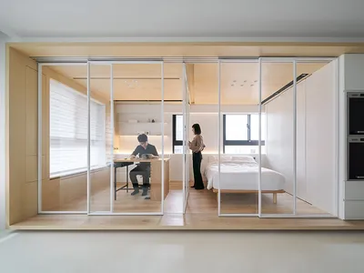 A minimalist bedroom with wood finishes and metal accents, featuring a white bed and a person standing near a window.