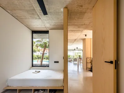 A minimalist bedroom with concrete flooring, a bed platform, and a large window overlooking a pool and palm trees.