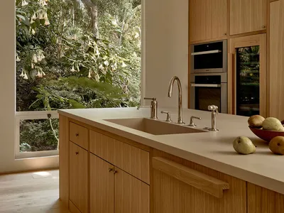 A modern kitchen with wooden cabinets, a sink, and fruits on the countertop, overlooking a lush green outdoor scenery through