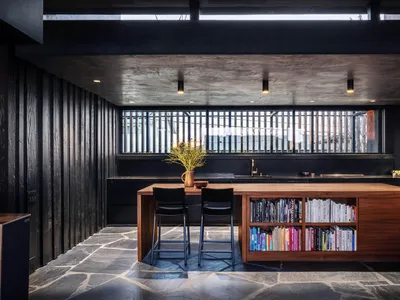 Modern minimalist kitchen with wooden island and stone flooring, featuring a bookshelf and stools.