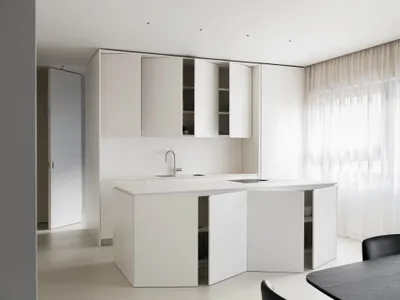 Modern, minimalist kitchen with white cabinets and countertops, featuring a sleek design and a small window.
