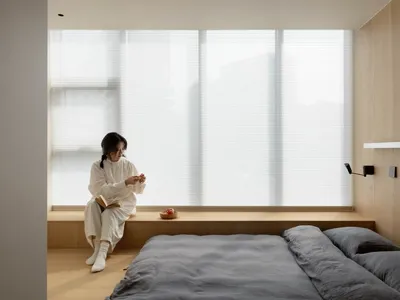 A minimalist bedroom featuring wooden elements with a person sitting by the window.