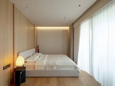 A minimalist bedroom featuring a white bed with a beige headboard, hardwood flooring, and lit floor lamp with white shade nex