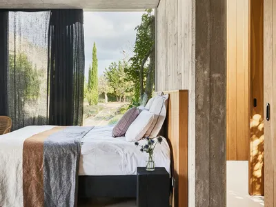 A minimalist bedroom with concrete walls, a wooden bed frame, and a large window overlooking a garden.