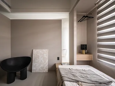 A minimalist bedroom featuring neutral-colored walls, a black chair, a bed with neutral bedding, and a wooden nightstand.