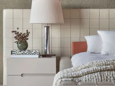 A modern bedroom featuring a leather headboard with a silver lamp and a blanket on the bed, set against a neutral-colored wal