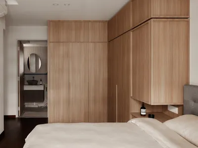 A minimalist bedroom featuring wooden cabinets, a white quilt, and a view into the attached bathroom with a circular mirror a