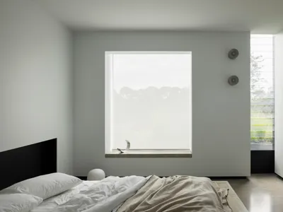 A minimalist bedroom with a concrete floor and walls, featuring a large window with a view of greenery.
