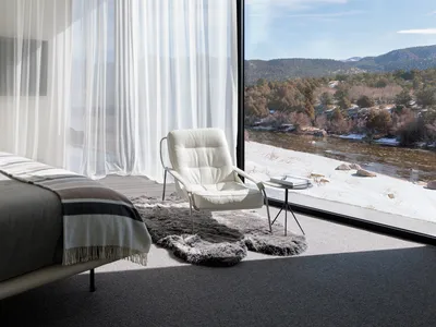 Modern minimalist bedroom featuring a white leather chair with a glass window overlooking mountains and a river.