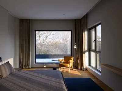 A modern bedroom featuring gray flooring and a blue rug, with natural light streaming through large windows and a sitting are