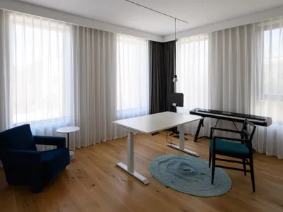 A modern home office with white curtains, wooden floors, and minimalist furniture.