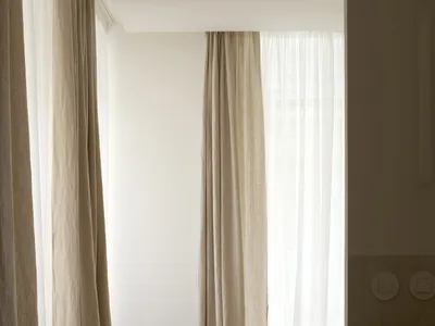 A minimalist bedroom with neutral-toned curtains, a single bed, and wooden flooring.