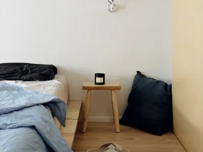 Minimalist bedroom with a bed, stool, and slippers on a wooden floor, featuring a wall-mounted light fixture.