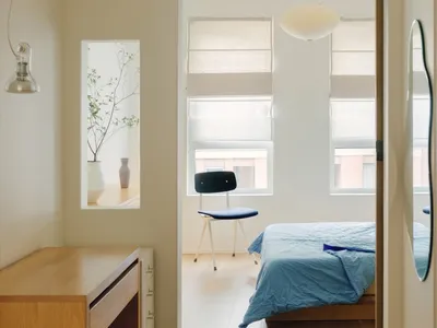 A modern, minimalist bedroom featuring hardwood flooring, a blue bedspread, and a dark blue upholstered stool.