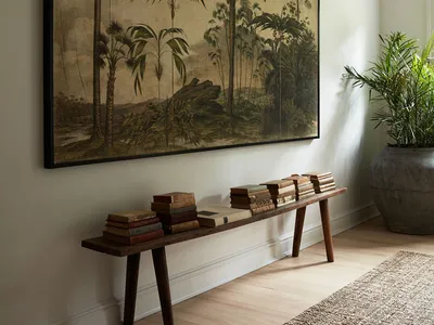 A photograph of a living room featuring a large framed painting of tropical foliage, a wooden bench adorned with stacked book