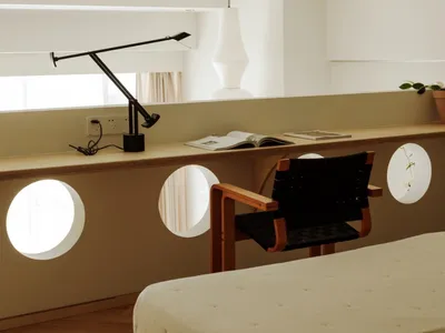 A minimalist bedroom featuring a simple bed with a wooden frame, a black leather armchair, and a floating desk with a black l